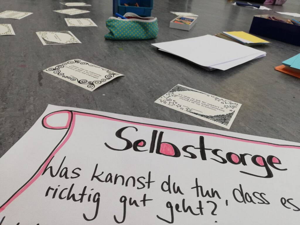 Ein Foto von dem Boden eines Seminarraums. Im Vordergrund liegt ein selbst geschriebener Zettel: "Selbstsorge. Was kannst du tun, dass es richtig gut geht?" Dahinter Affirmationskarten, Papier und Stife.